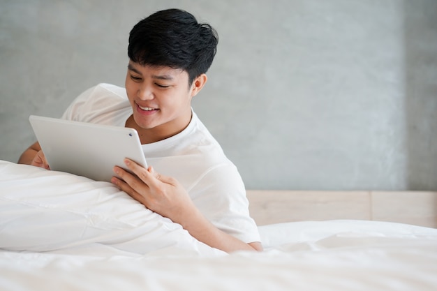 Hombre asiático joven jugando tablet en cama en fin de semana