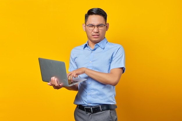 Hombre asiático joven pensativo asiático con gafas usando una computadora portátil aislada en un concepto de empresario y emprendedor de fondo amarillo