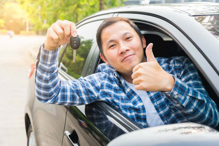 Hombre asiático joven que muestra el pulgar para arriba y la mano que lleva a cabo su llave en el coche. Concepto de alquiler de coche o compra de coche.
