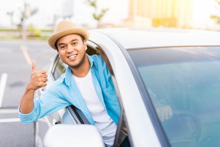 Hombre asiático joven que sonríe y que muestra el pulgar para arriba en su coche.
