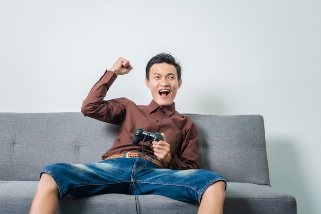 Hombre asiático joven que sostiene la palanca de mando para jugar al videojuego del fútbol mientras que se sienta en el sofá en sala de estar.