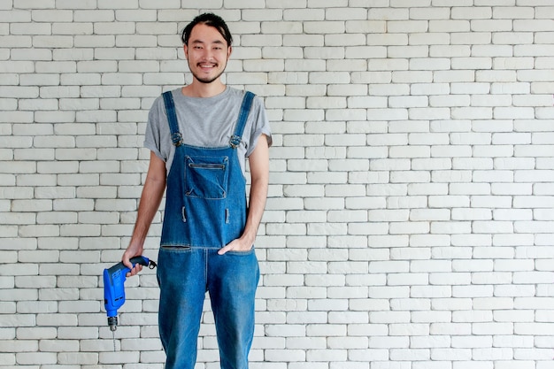 Hombre asiático joven que sostiene el taladro eléctrico de pie delante de la pared de ladrillo blanco, sonriendo y mirando a la cámara, concepto de bricolaje en casa