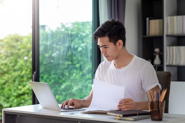 Hombre asiático joven que trabaja en una computadora portátil con un documento en casa.