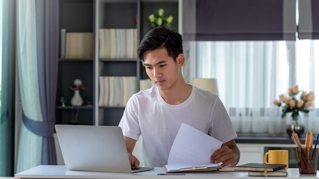 Hombre asiático joven que trabaja en una computadora portátil con un documento en casa.