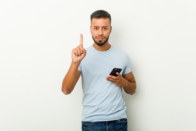 Hombre asiático joven de raza mixta sosteniendo un teléfono mostrando el número uno con el dedo.