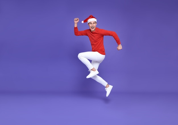 Hombre asiático joven en traje casual rojo con sombrero de Santa saltando y cara de sonrisa en la pared púrpura. Feliz año nuevo concepto.