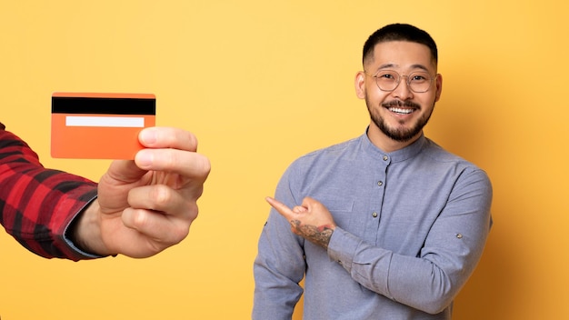 Un hombre asiático maduro y alegre señalando con el dedo la mano con una tarjeta de crédito aislada en el fondo amarillo del estudio