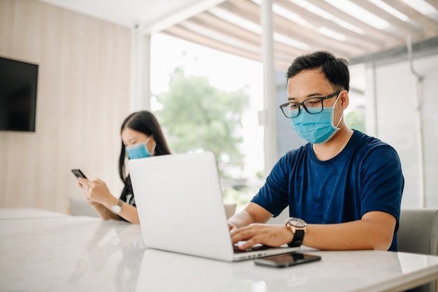 Hombre asiático con una máscara y usando una computadora portátil en la oficina o en el aula para la prevención del virus COVID-19.