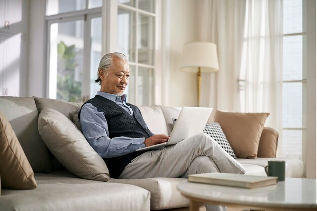 hombre asiático mayor sentado en el sofá mirando una computadora portátil