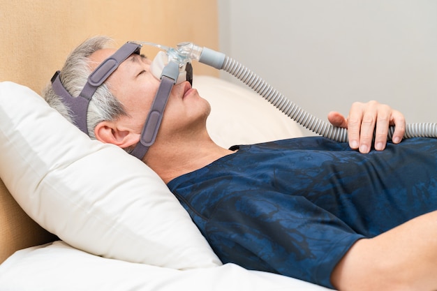 Hombre Asiático Con La Apnea Del Sueño Usando La Máquina De CPAP, Usando La  Máscara De Arnés Que Conecta Al Tubo De Aire, Enfoque Selectivo En El  Hombre Máscara Y El Arnés