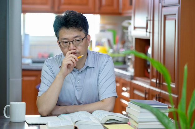 Hombre asiático de mediana edad sentado en el escritorio en casa, leyendo un libro y estudiando.