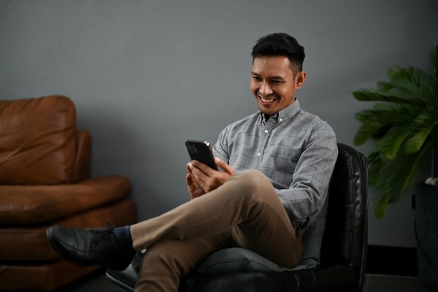 Un hombre asiático milenario feliz que usa su teléfono inteligente mientras se relaja en la sala de estar