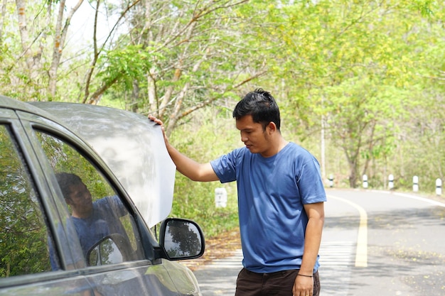 Hombre asiático mira el motor de un auto roto mientras viaja