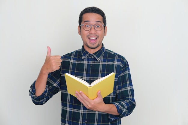 Hombre asiático mirando la cámara y levantando el pulgar emocionado al sostener un libro