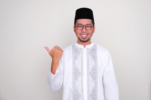 Hombre asiático musulmán sonriendo a la cámara y apuntando hacia el lado derecho con el pulgar