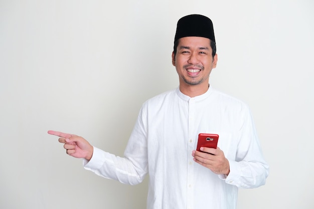 Hombre asiático musulmán sonriendo feliz mientras sostiene el teléfono móvil y señala hacia el lado derecho