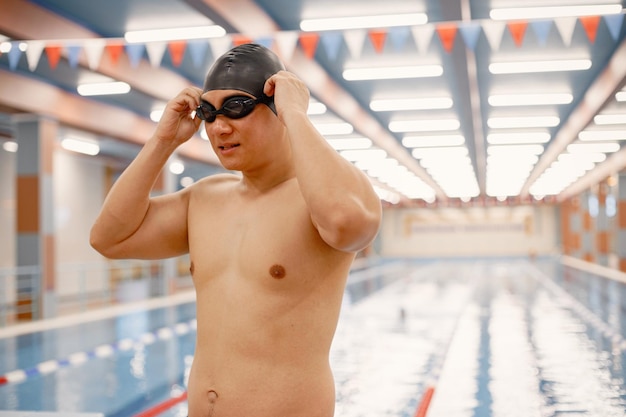 Hombre asiático parado en una piscina cubierta y con sombrero y gafas