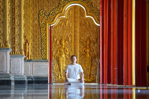 Hombre asiático de pelo largo relaja la meditación con todo traje blanco sentado frente al papel tapiz dorado de budista en el templo, Tailandia.