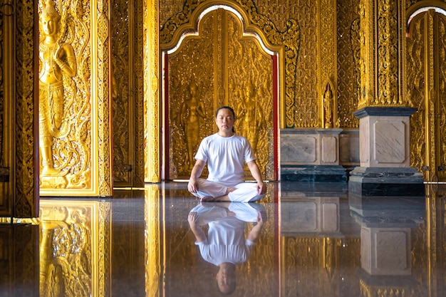 Hombre asiático de pelo largo relaja la meditación con todo traje blanco sentado frente al papel tapiz dorado de budista en el templo, Tailandia.