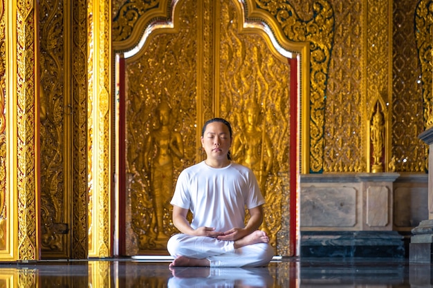 Hombre asiático de pelo largo relaja la meditación con todo traje blanco sentado frente al papel tapiz dorado de budista en el templo, Tailandia.