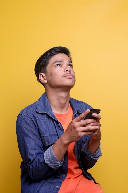 Un hombre asiático pensativo mirando hacia arriba y pensando mientras sostiene el teléfono móvil