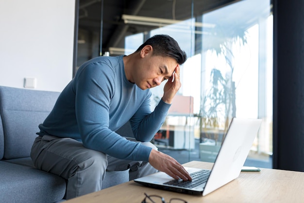 Hombre asiático pensativo que trabaja en la oficina en una computadora portátil hombre de negocios ansioso en la oficina moderna frustrado deprimido