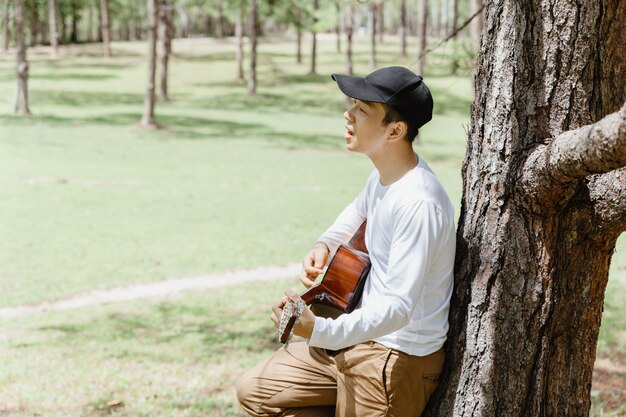 Hombre asiático de pie y cantando con la guitarra bajo el árbol de pino.