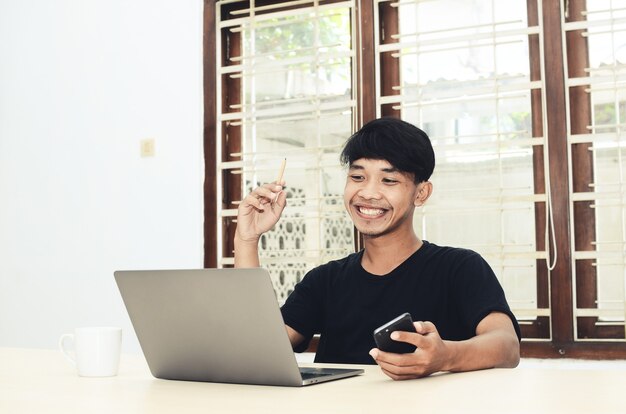 El hombre asiático que está sentado en la mesa de la oficina con una camisa negra está sonriendo frente a la computadora portátil