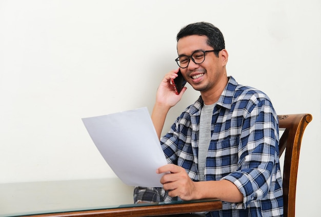 Hombre asiático que muestra una expresión feliz cuando está en una llamada telefónica y lee una carta