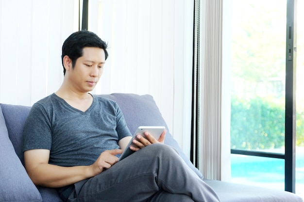 Hombre asiático que se sienta en el sofá y que usa la tableta en casa, estilo de vida, comunicación de la tecnología