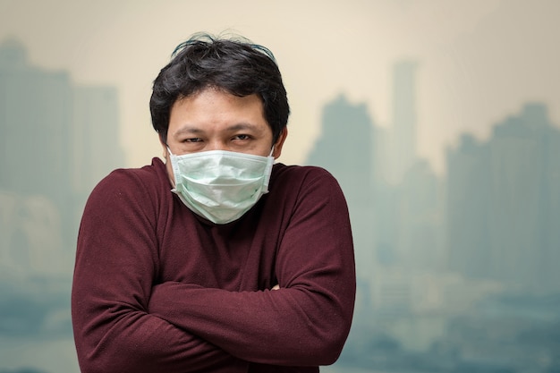 Hombre asiático que usa la mascarilla contra la contaminación del aire con frío en el balcón de High Apartment