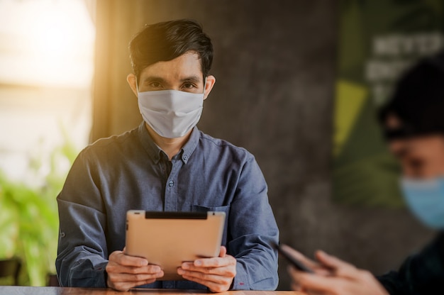 Hombre asiático que usa una tableta en línea trabajando con una máscara facial que protege el estilo de vida del virus corona nuevo normal, el hombre vive en la cafetería interior