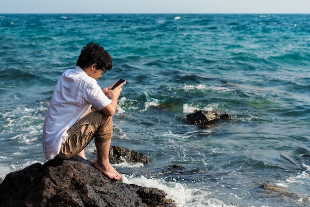 Hombre asiático que usa el teléfono que mecanografía el mensaje a la charla en red social mientras que se sienta en roca en el mar.