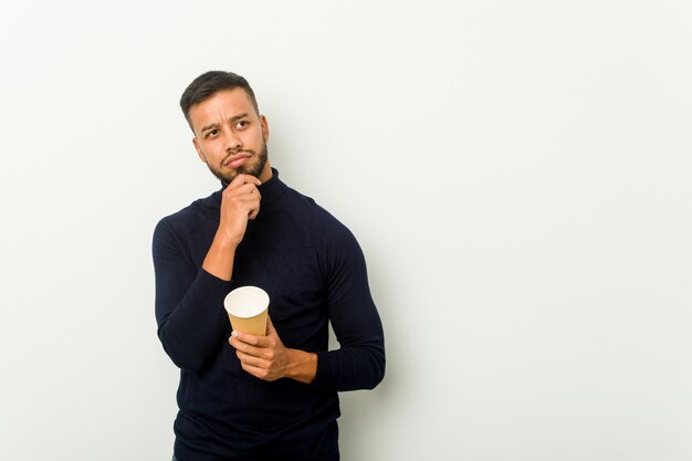 Hombre asiático de raza mixta joven sosteniendo un café para llevar mirando hacia los lados con expresión dudosa y escéptica.