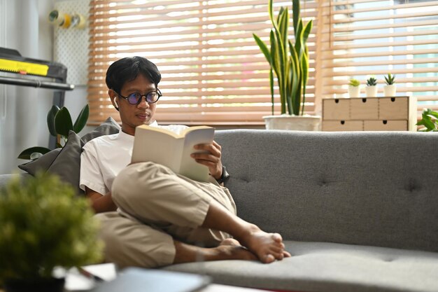 Hombre asiático relajado sentado en el sofá en casa y leyendo un libro el fin de semana