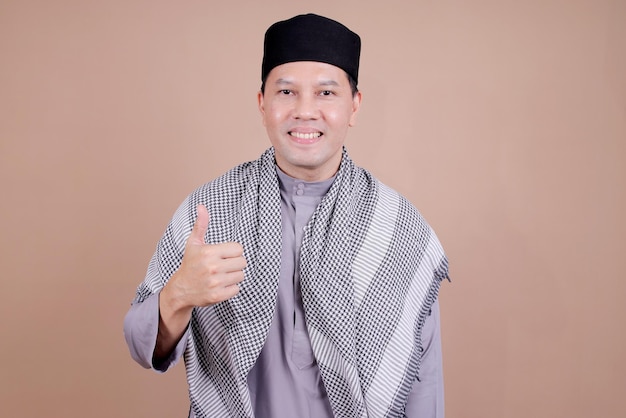 Hombre asiático con ropa musulmana sonriendo levantando el pulgar para la publicidad sobre un fondo beige