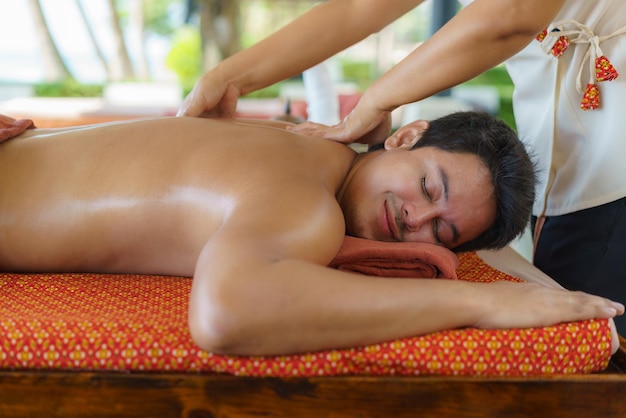 Hombre asiático en el salón de spa de playa recibiendo masaje con aceite cerca de la playa y mirando feliz en Tailandia