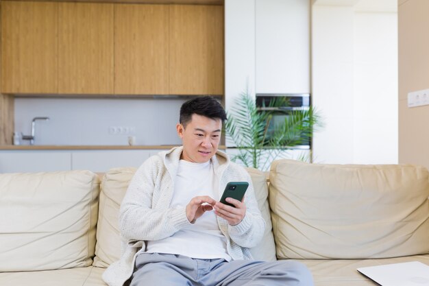 Hombre asiático sentado en casa en el sofá, usando un teléfono inteligente, serio escribiendo un mensaje en el teléfono