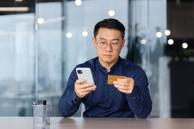 Hombre asiático serio y pensante dentro de la oficina usando la aplicación en el teléfono y la tarjeta de crédito bancaria para la tienda en línea