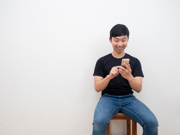 Hombre asiático con smartphone en la mano sentado en la silla cara de sonrisa feliz sobre fondo de pared blanca