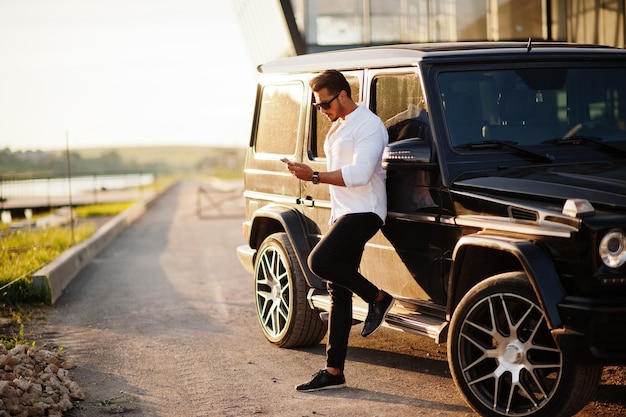 Un hombre asiático sólido con camisa blanca y gafas de sol posó cerca de un camión suv de la mafia negra con teléfono móvil.