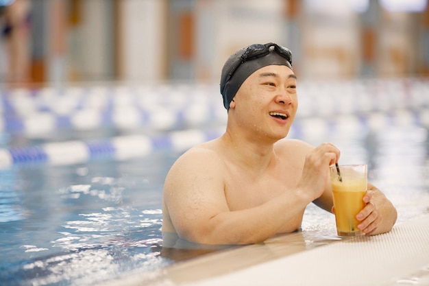 Hombre asiático con sombrero y gafas bebiendo un jugo de naranja en la piscina