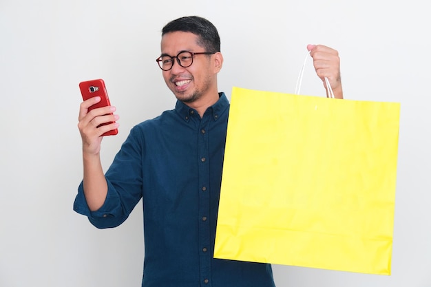 Hombre asiático sonriendo feliz cuando mira su teléfono mientras muestra una gran bolsa de compras amarilla