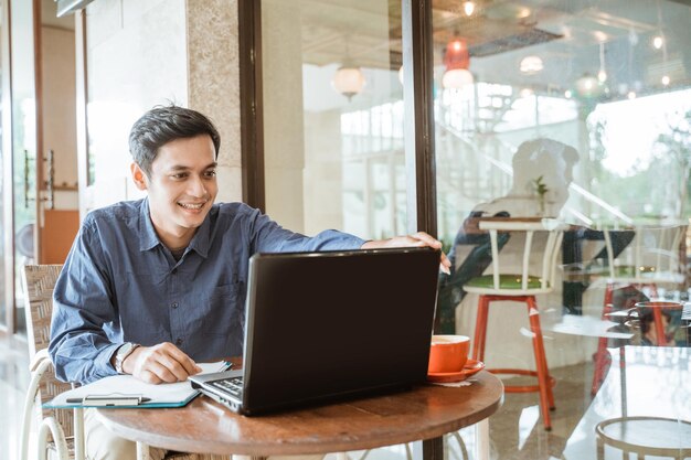 Foto hombre asiático sonriendo mientras configura una computadora portátil antes de una conferencia en línea mientras trabaja en un espacio de trabajo conjunto