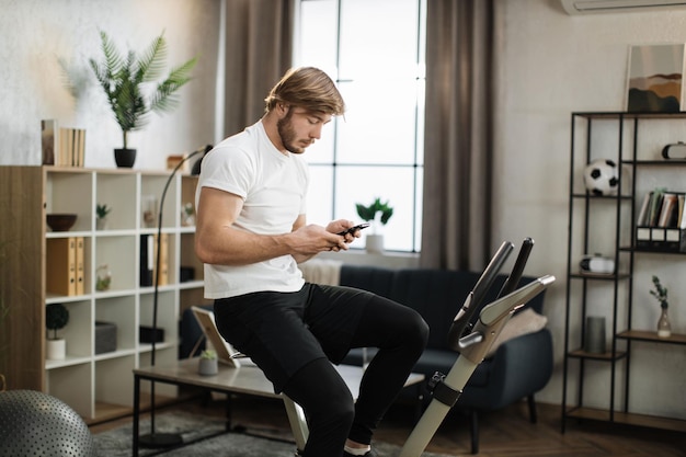 Hombre asiático sonriente en forma saludable entrenando en casa en bicicleta estática durante el ejercicio sosteniendo el teléfono