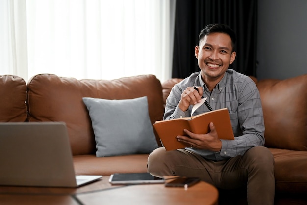 Hombre asiático sonriente haciendo una lista o tomando notas en su cuaderno mientras se sienta en el sofá