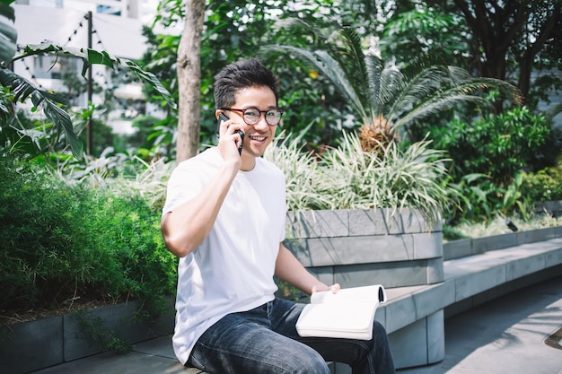 Hombre asiático sonriente leyendo un libro y hablando por teléfono inteligente