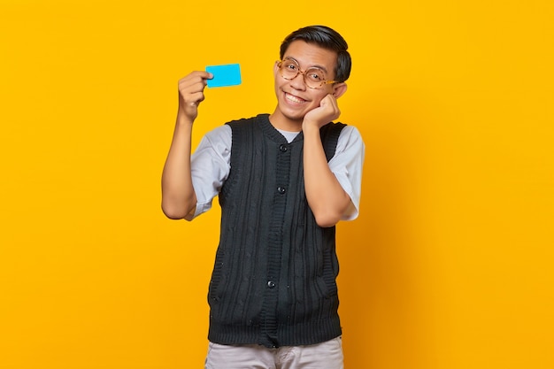 Hombre asiático sonriente que sostiene la tarjeta de crédito con la boca abierta y las manos en las mejillas sobre fondo amarillo