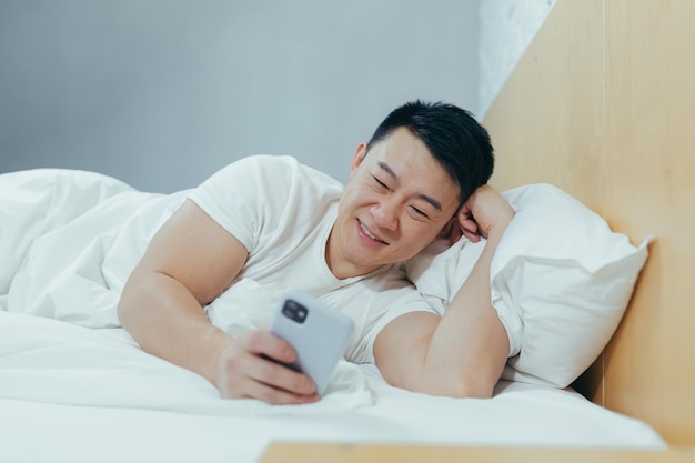 Hombre asiático sonriente y soñoliento acostado en la cama bajo una manta mirando a la cámara y sonriendo usando el teléfono leyendo las noticias de la mañana en línea