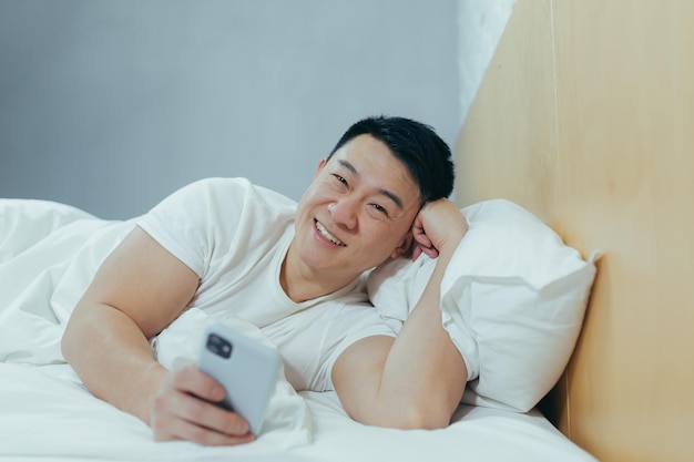 Hombre asiático sonriente y soñoliento acostado en la cama bajo una manta mirando a la cámara y sonriendo usando el teléfono leyendo las noticias de la mañana en línea